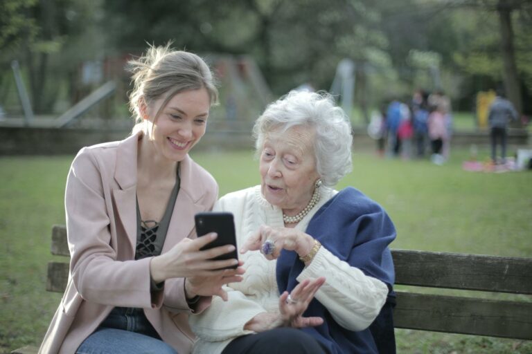 Mother supporting grandmother with dementia, on wooden park bench, browsing mobile phone